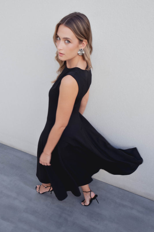 Vestido midi negro de cuello redondo y corte sisa. Bajo con paneles o nesgas que dan vuelo. Cierre con cremallera en la espalda.